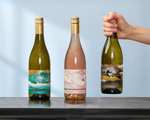 Three bottles of white wine on a table, with one being picked up showing a white person's hand and wrist.